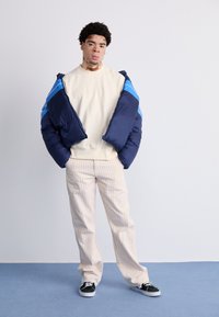 Tengerészkék és kék puffertakaró kabát, alatta krémszínű sweatshirt, csíkos bézs nadrág és fekete sportcipő. Egyszínű háttér előtt.