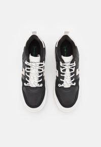 Baskets en cuir noir avec des accents blancs, surface texturée et détails perforés. Comportent des lacets blancs et un logo vert sur la semelle intérieure.