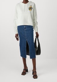 Sudadera blanca corta con un logotipo gráfico, combinada con una falda de mezclilla con una abertura frontal. Se ve un bolso negro y sandalias de tacón cuadrado.