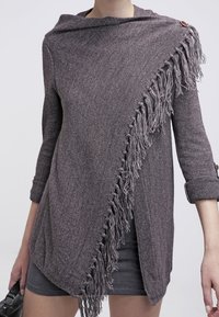 Maglione grigio testurizzato con cappuccio, caratterizzato da un dettaglio di frange diagonali su un lato e un accredito con bottone sul colletto.