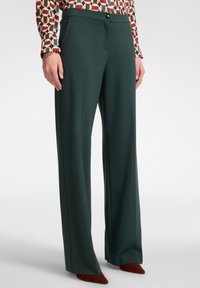 Donna che indossa pantaloni larghi verde scuro, una blusa a maniche lunghe a fantasia, un bracciale dorato e scarpe marroni a punta.