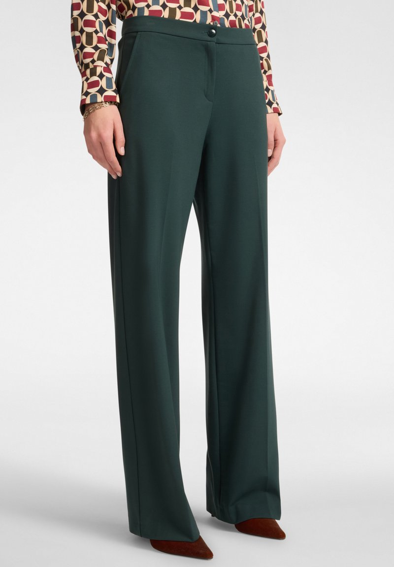 Donna che indossa pantaloni larghi verde scuro, una blusa a maniche lunghe a fantasia, un bracciale dorato e scarpe marroni a punta.
