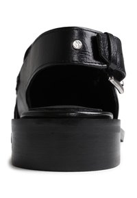 Sandalia de cuero negro con correa ajustable, tacón cuadrado y detalle de hardware plateado. Textura suave con un diseño de espalda abierta.