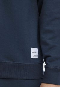 Temno modra pulover s ribanim manšetom in spodnjim robom. Ima belo oznako na spodnjem robu, ki pravi "BLEND DEDICATED TO DENIM PERFECTION."