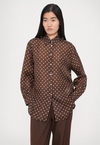 Cămașă maro cu buline, realizată dintr-un material ușor, cu nasturi în față, mâneci lungi și guler clasic, asortată cu pantaloni maro largi.