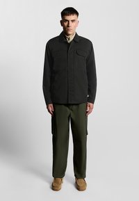 Mørkegrøn skjortejakke med to frontlommer, parret med olivengrønne cargo-bukser og lysebrune slip-on sko. Enkel design og funktionalitet.