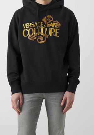 Osoba nosząca czarną bluzę z kapturem z złotym tekstem "Versace Jeans Couture" i dekoracyjnymi zawijasami, w połączeniu z jasnoszarymi dżinsami.