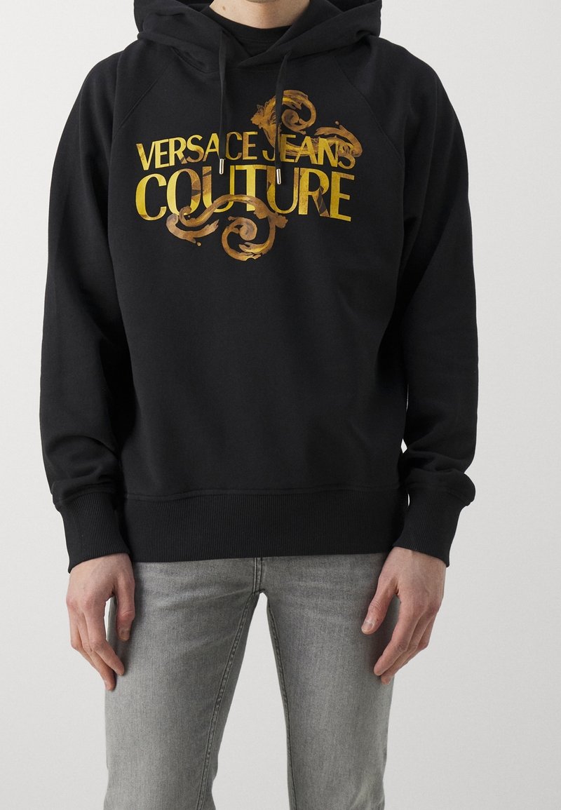 Personne portant un sweat à capuche noir avec un texte doré "Versace Jeans Couture" et des volutes décoratives, associé à un jean gris clair.