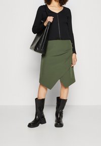 Top noir à manches longues côtelé avec fermeture éclair à l'avant ; jupe asymétrique vert olive avec boutons sur le côté ; bottines noires ; sac fourre-tout noir.