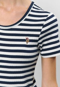 Jūras spēku un baltas svītrām raustītaad t-krekls ar ovālas formas dekoltē un īsām piedurknēm; tam ir mazs izšūts attēls pie apkakles.