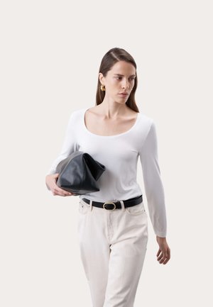 Frau mit langen braunen Haaren, die ein weißes Oberteil und helle Hosen trägt, hält eine große schwarze Clutch und blickt seitlich vor neutralem Hintergrund.