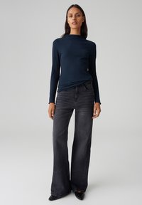 Marineblaues, geripptes Langarmoberteil mit hohem Kragen, kombiniert mit schwarzen, weiten Jeans und spitzen, schwarzen Schuhen, um eine elegante, moderne Silhouette zu erzeugen.