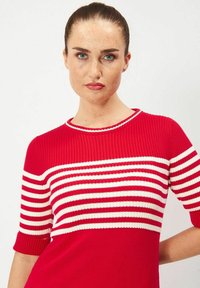 Punainen neulottu villapaita lyhyillä hihoilla, jossa on vaaleita vaakasuoria raitoja yläosassa ja yhtenäinen punainen alaosa.