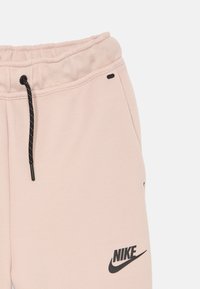 Ljust rosa Nike sweatpants med svart dragsko och svart Nike-logga på låret, visade från midjan till mitten av benet.