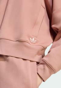 Svijetloroze dukserica s krojenim dizajnom, koja ima zatvaranje na zip i mali bijeli Adidas Trefoil logo na lijevom džepu.