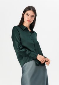 Camicia di raso verde scuro con collo, maniche lunghe e chiusura frontale con bottoni, abbinata a una gonna di raso grigia.