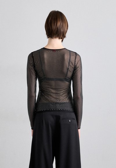 Schwarzes, durchsichtiges Oberteil mit langen Ärmeln und weißen Punkten, das über einen runden Ausschnitt und ein figurbetontes Design verfügt. Kombiniert mit schwarzen, hochgeschnittenen Hosen.