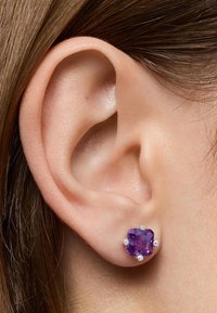 Orecchini in pietra preziosa viola incastonati in quattro griffe d'argento, indossati su un lobo dell'orecchio di un modello, con pelle liscia e capelli castani lisci sullo sfondo.