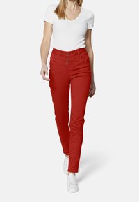 Pantalones altos rojos hechos de una tela elástica, con tres botones en la parte delantera, un corte ajustado y bolsillos laterales. Combinados con una blusa blanca y zapatillas.