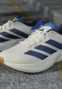 Chaussures de sport blanches avec des rayures bleu marine et des accents bleus, lacées et posées sur une surface à texture rugueuse.