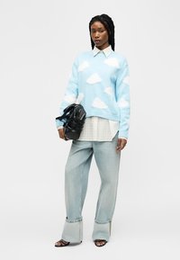 Femme avec des cheveux tressés portant un pull bleu orné de nuages blancs, une chemise blanche à col, un jean à jambes larges, des sandales noires, et tenant un sac noir.