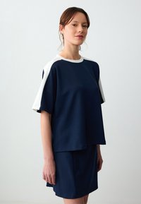 Top blu navy a maniche corte con colletto e dettagli bianchi, caratterizzato da una vestibilità morbida e una texture liscia. Indossato con una gonna blu navy coordinata.
