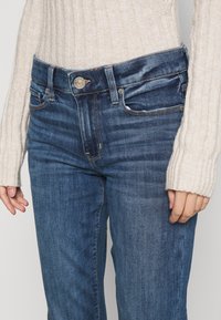 Džínové kalhoty v tmavě modré s vybledlou texturou, designem s pěti kapsami a knoflíkovým zapínáním v pase. Spárované s krémovým pleteným svetrem.