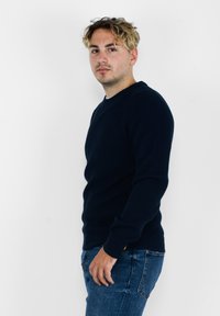 Maglione a coste blu navy con maniche lunghe e scollatura tonda, abbinato a jeans in denim blu. Il maglione presenta un tessuto texturizzato e una decorazione minimale.