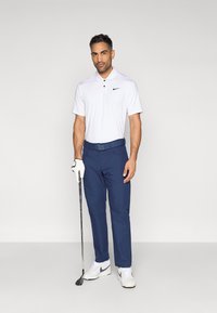 Λευκό πόλο πουκάμισο με γιακά και μαύρο λογότυπο, σε συνδυασμό με μπλε παντελόνια και λευκά αθλητικά παπούτσια. Κρατάει ένα γκολφ ρόπαλο, φοράει λευκό γάντι.