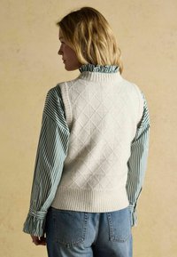 Eine Frau mit schulterlangem blonden Haar trägt eine weiße, diamantmusterte Strickweste über einer grün-weiß gestreiften Bluse und blauen Jeans und schaut nach links.