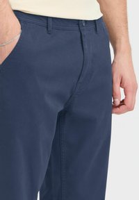 Morske bombažne hlače z ravnim krojem, stranskimi žepi in pasom s peteljkami, vidnimi šivi in gladko teksturo.