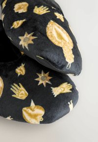 Zapatillas de tela negras con una superficie suave y texturizada, decoradas con patrones de manos, soles y manzanas en amarillo. Diseño de puntera redondeada y bordes acolchados.