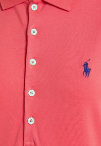 Czerwona koszulka polo z teksturowanej tkaniny, z trzema guzikami przy kołnierzyku i jasnoniebieskimi guzikami. Wyhaftowane ciemnoniebieskie logo na lewej piersi.