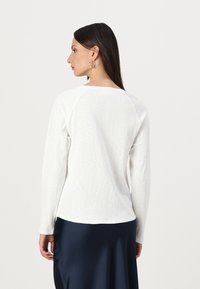 Long-sleeve white top made of lightweight fabric with a textured finish and a rounded hem, worn with a blue skirt.