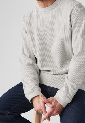 Mand iført en lysegrå struktureret sweater og mørkeblå jeans, siddende på en træskammel med hænderne foldet foran.