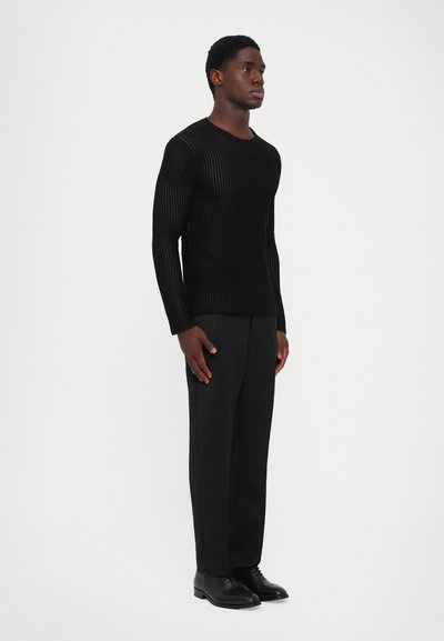 Άνδρας που στέκεται στο πλάι, φορώντας μαύρο ριγέ μακρυμάνικο πουκάμισο, μαύρο παντελόνι και μαύρες κοσμηματοκότες παπούτσια, απέναντι από ένα λευκό φόντο.