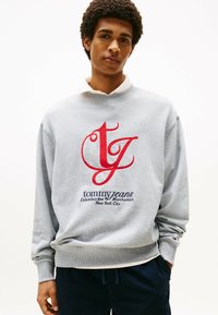 Sudadera gris con un logo bordado en rojo y azul marino. Puños y dobladillo acanalados, con un cuello sobre una camisa blanca debajo.