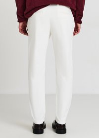 Pantaloni bianchi su misura con una finitura testurizzata, vestibilità slim e due tasche posteriori, abbinati a scarpe nere su uno sfondo semplice.