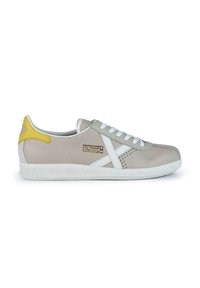 Zapatillas beige con cordones blancos, que presentan un acento en el talón amarillo y franjas laterales blancas distintivas. Textura de cuero suave con suela de goma.