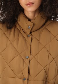 Gilet matelassé marron avec un col haut et des boutons-pression argentés. Le tissu présente une texture lisse et un motif en diamant sur l'ensemble.