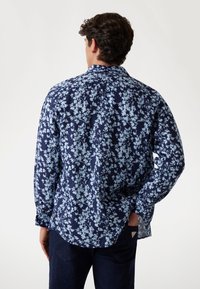 Camicia a maniche lunghe di colore blu scuro con motivo floreale blu chiaro. Caratterizzata da un colletto classico e polsini con bottone. Realizzata in tessuto traspirante.
