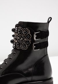 Stivaletto in pelle nera con un bordo decorativo di perline caratterizzato da grandi accenti neri e argentati, fasce a doppia fibbia e una finitura liscia.