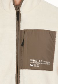 Fleecejacka med en krämfärgad kropp och en brun ficka som har texten "WHSTLR / OUTDOOR ULTIMATE PROTECTIVE WEAR." Dragkedjestängning.