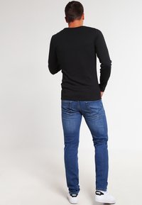 Man i svart långärmad skjorta, blå jeans och vita Nike-sneakers, som står med ryggen mot kameran framför en helt vit bakgrund.