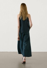 Haut sans manches vert foncé associé à un pantalon large, tous deux en tissu texturé. Le mannequin porte des sandales à lanières.