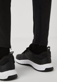 Zwarte sportieve schoenen met een gestructureerde bovenkant, witte middenzool en zwarte buitenzool; gecombineerd met zwarte tappered broek met ritsdetails.