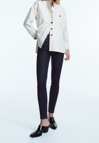 Kremowa koszula typu button-up z zielonymi guzikami, ciemne wąskie dżinsy oraz błyszczące czarne mule na obcasie, podkreślające teksturę i kontrast kolorów.