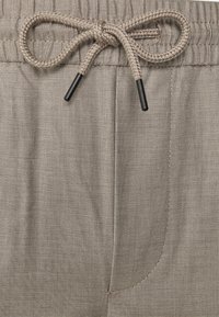 Pantalones de tela beige texturizada con cintura elástica y cordón trenzado atado con puntas de plástico negro.
