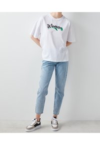 Valkoinen lyhythihainen t-paita, jossa lukee "Nainen" vihreällä ja mustalla. Yhdistetty vaaleansinisiin kapeisiin farkkuihin ja erottuviin mustavalkoisiin lenkkareihin.