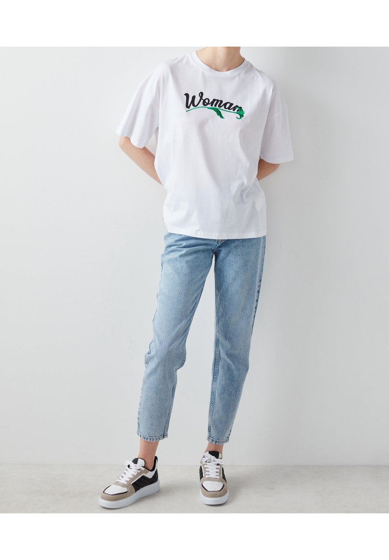 Bílý krátký rukáv tričko s nápisem "Žena" tištěným zelenou a černou barvou. Kombinováno se světle modrými skinny džíny a výraznými černobílými teniskami.